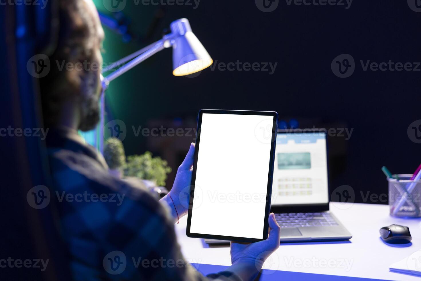 Man vertically holding tablet with isolated white screen in front of his laptop. Male freelancer working from home, focused on digital device displaying blank chromakey mockup template. photo