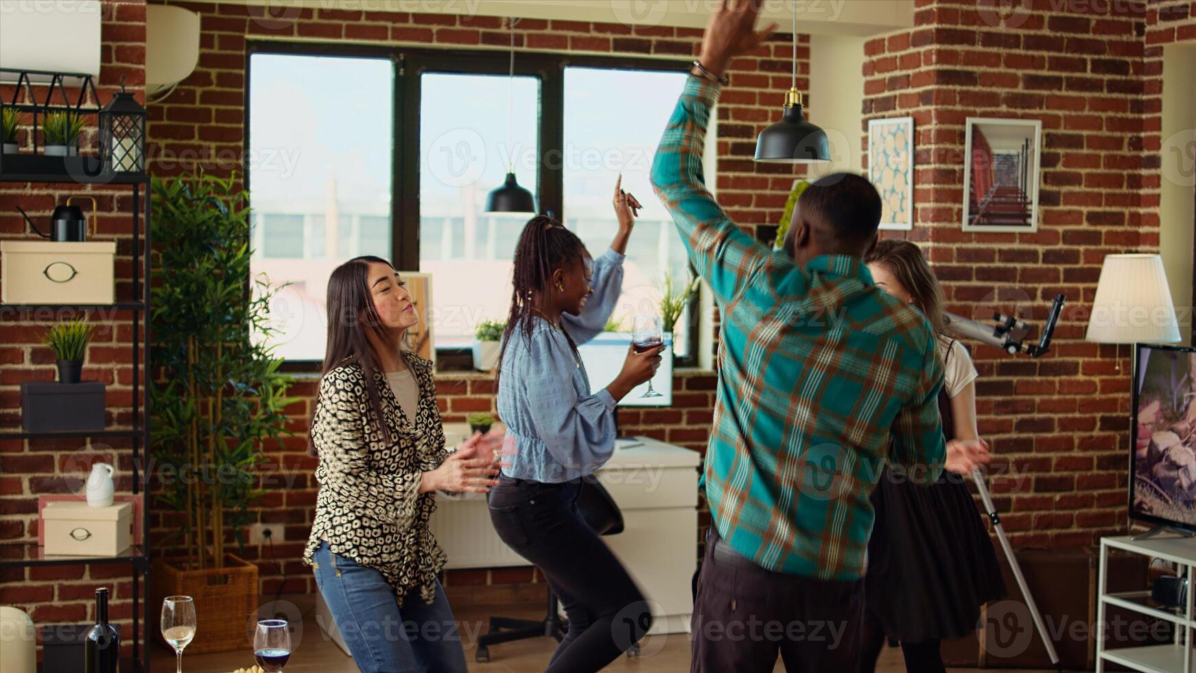 Multicultural youthful group of adults dancing at apartment party while drinking wine and champagne. Friends jumping around house, busting dance moves to energetic music in cozy living room photo