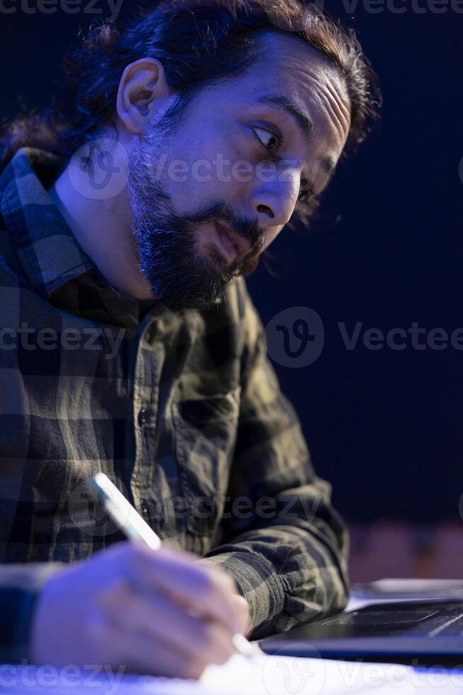 Sitting in his apartment, focused university student working diligently on his assignment, switching between his laptop and handwritten notes. Closeup of man writing on his notebook. photo