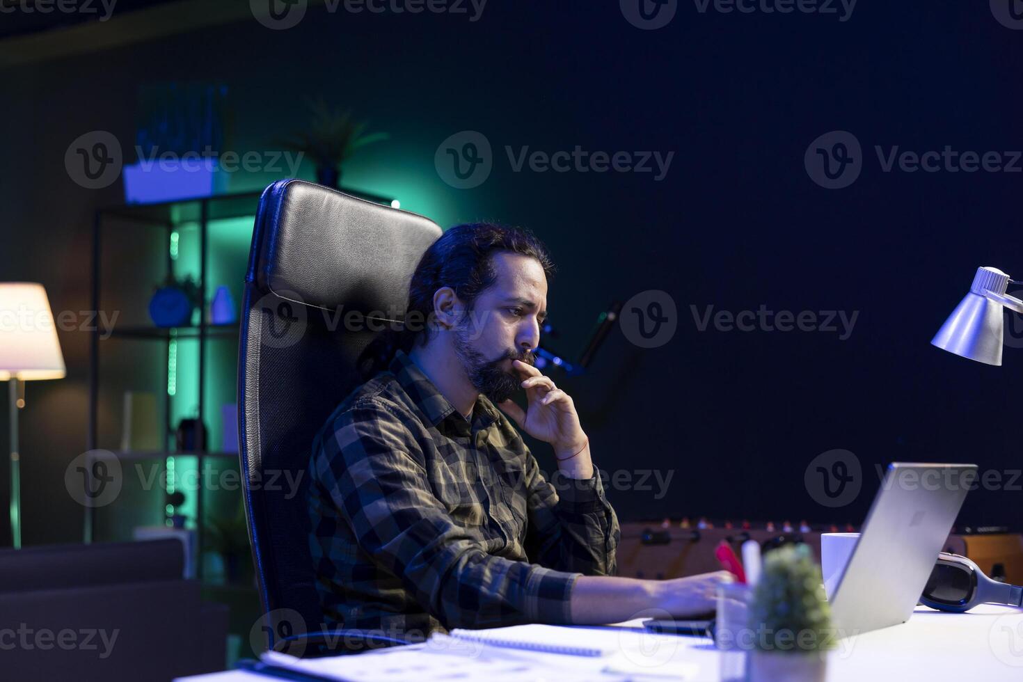 Concentrated blogger sitting at a table, working remotely from home. With laptop and focused expression, he is reviewing and typing, engaged in the creative process of blogging. photo