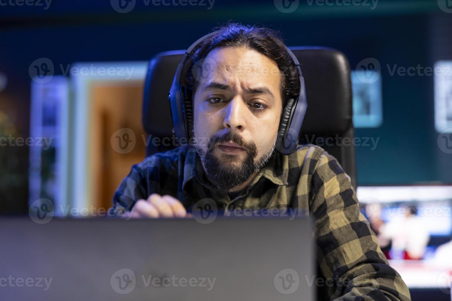 Close up of serious man wearing headphones and looking at his laptop. Male individual working from home, using wireless headset for music while browsing the internet on device. photo