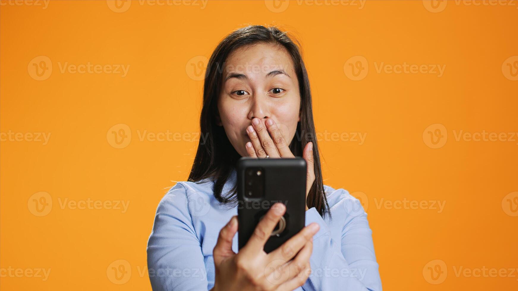 Asian woman answers online videocall in studio, talking to people on internet remote connection. Young adult smiling and waving at phone screen, online videoconference meeting over background. photo