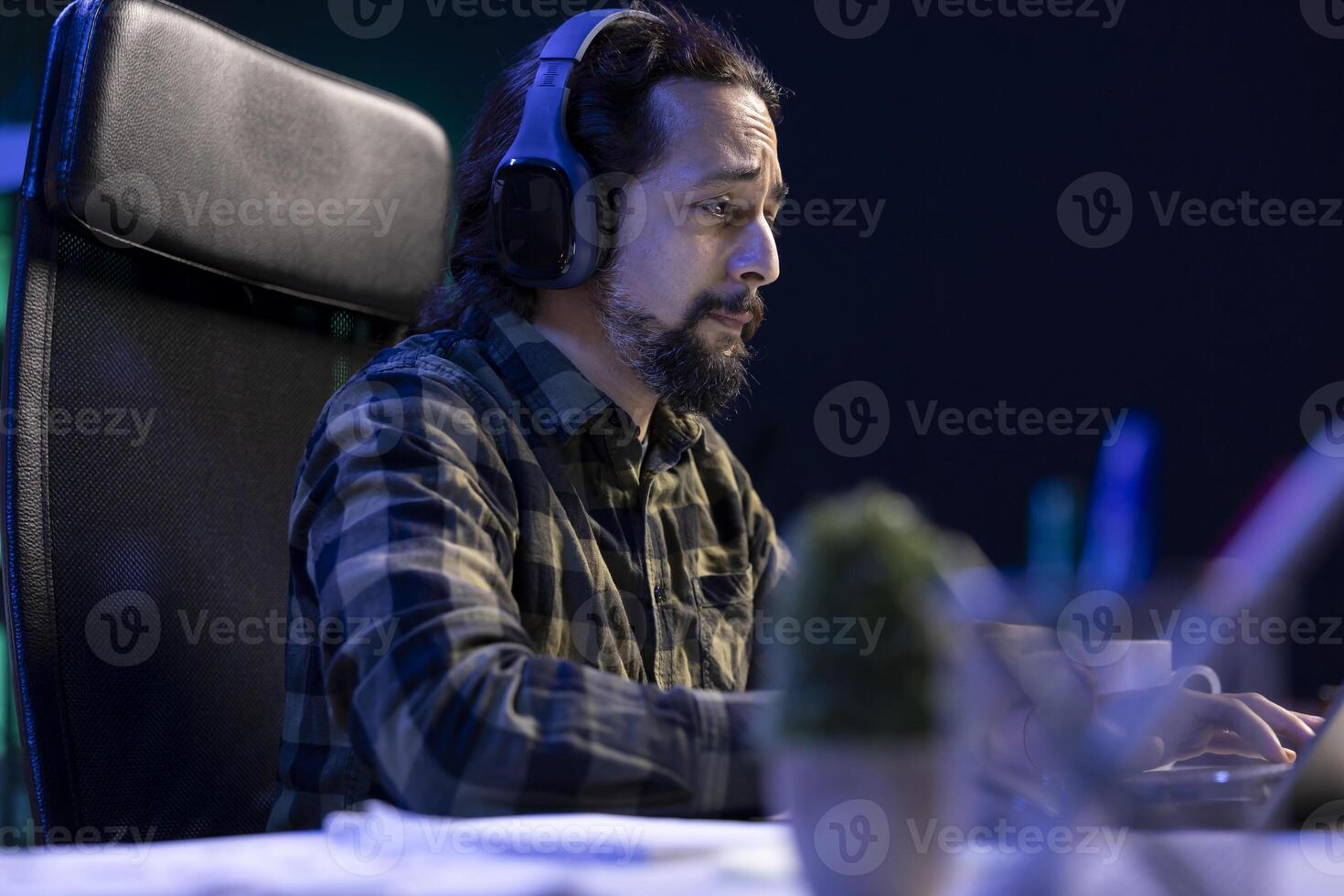 A focused man wearing a wireless headset attends a video call meeting with clients on his portable computer. A male with a sad expression works remotely, using a laptop and headphones. photo
