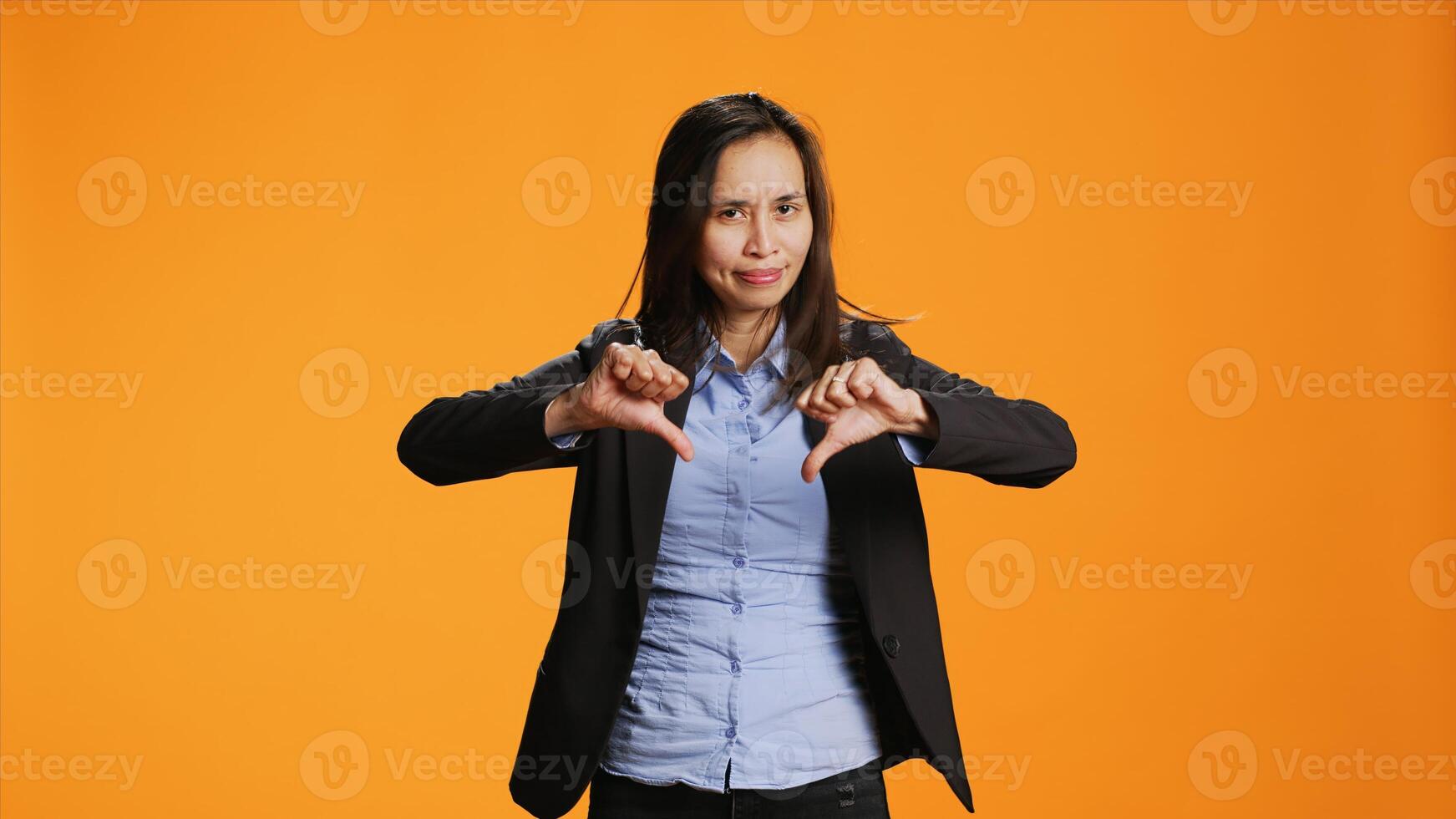 Asian employee showing dislike sign in front of camera, presenting thumbs down with unhappy facial expressions. Young adult does rejection and disagreement symbol over orange backdrop. photo