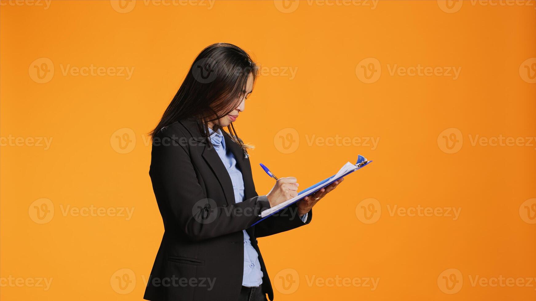 Asian manager writes important data on papers with clipboard, taking notes to create full report in front of orange background. Young woman in business clothing filling in documents, keeping track. photo