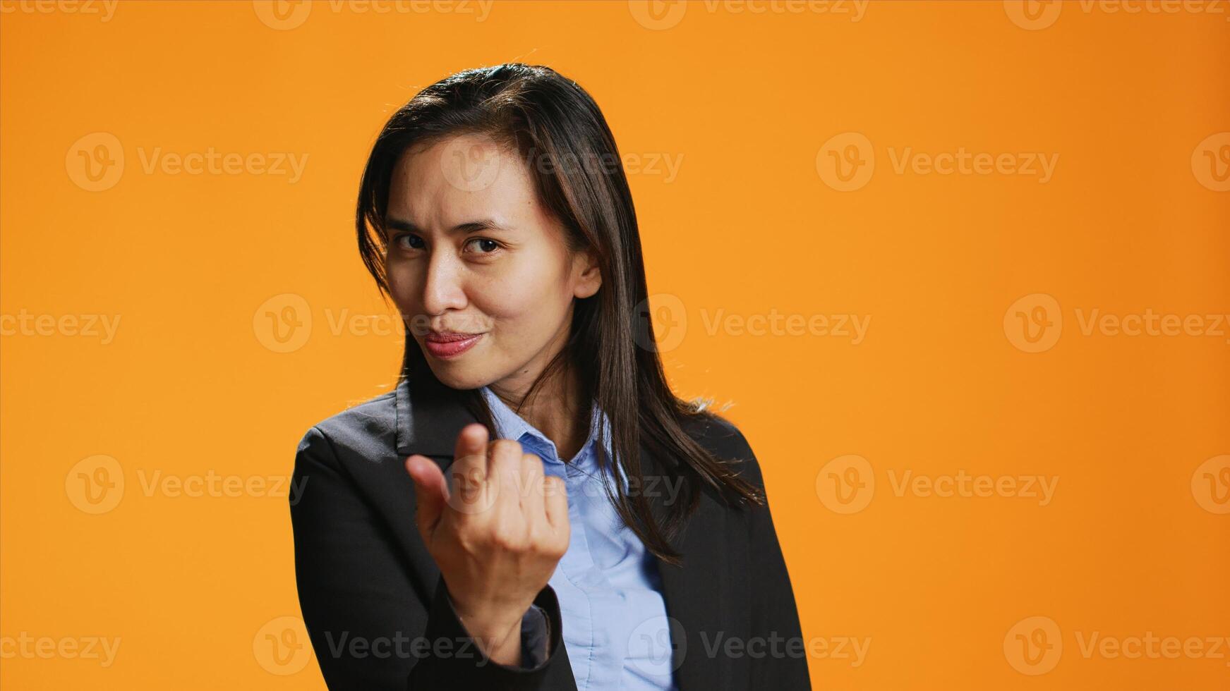 Female model calling for someone on camera, doing hailing gesture against orange background. Asian woman with office clothing inviting person to come closer, friendly adult. photo