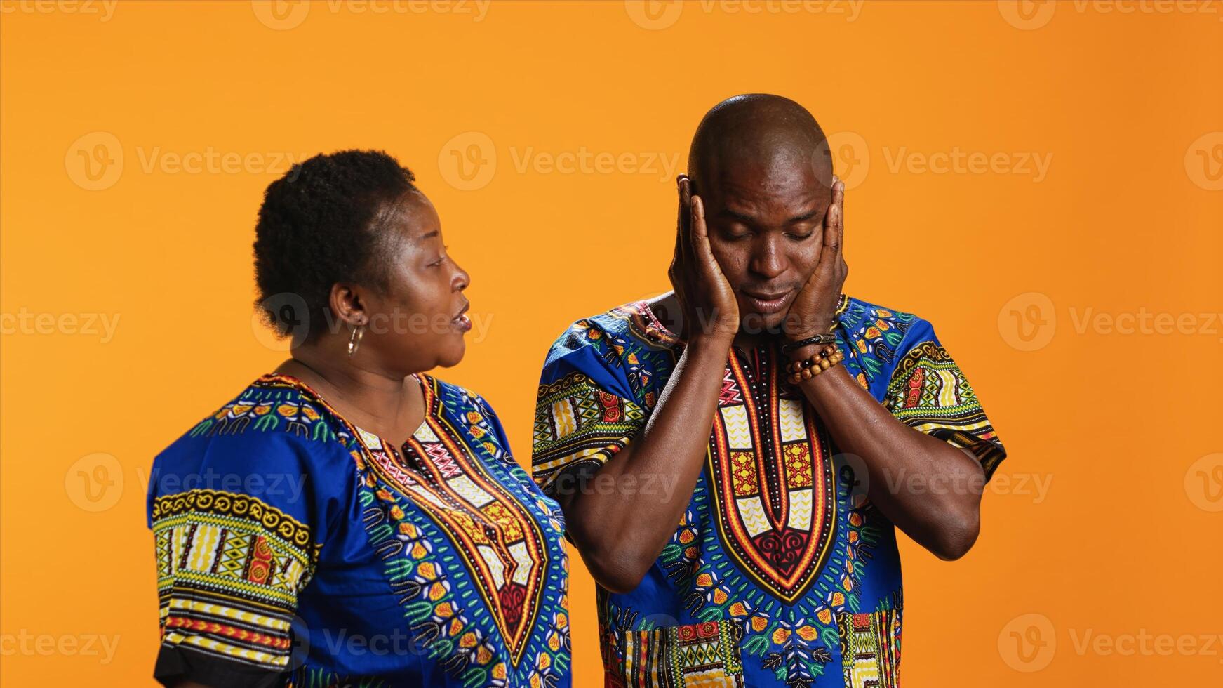 Angry wife yelling at her husband and starting fight, having argument over orange background. Tired man covering his ears and trying to ignore his partners screaming in studio. photo