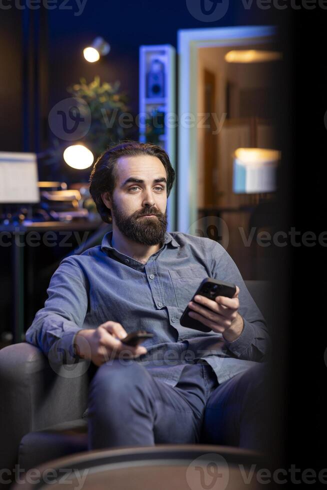 Man holding remote control while watching interesting movie on streaming service, enjoying time off from work. Person on couch surprised by high quality film broadcasted on TV photo