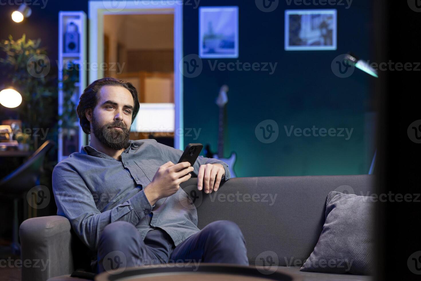 Man lounged on couch texting friend on smartphone, inviting him to watch movie on streaming service. Person at home discussing with mate on mobile phone, recommending TV show photo