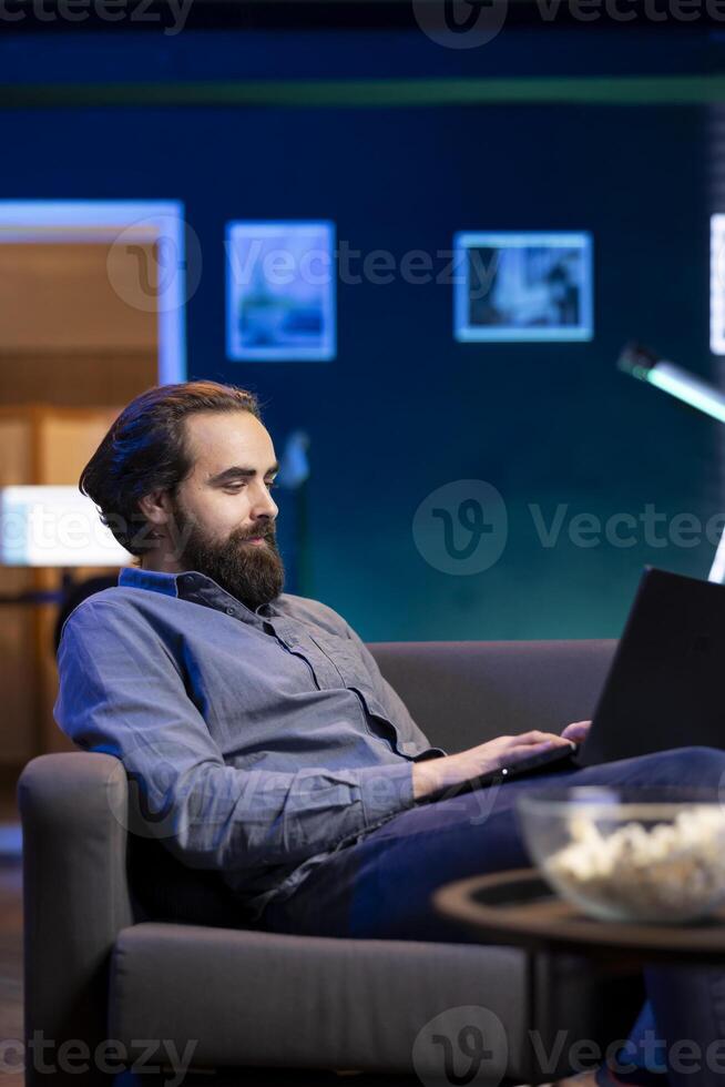 Smiling man enjoying bowl of popcorn while watching TV and solving tasks on laptop. Spectator on couch in living room eats snacks during movie and scrolls on social media using notebook photo
