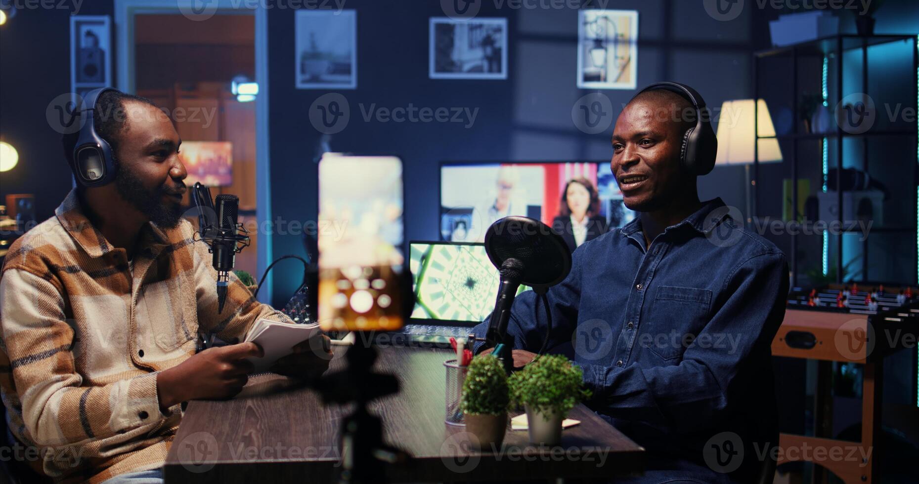 Man on podcast filming with cellphone on tripod while talking with guest during marathon stream for humanitarian cause, panning shot, gathering donations from viewers, handheld camera shot photo