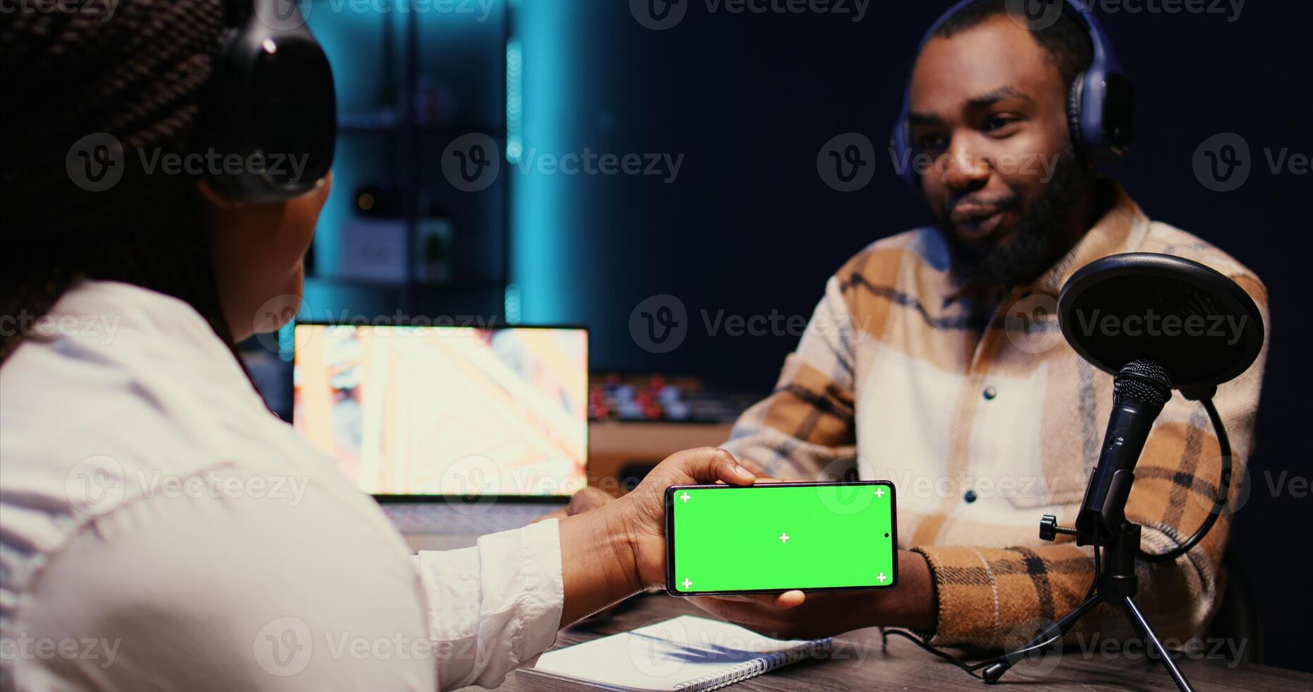 Man invited to podcast watching internet clips on green screen smartphone with host in studio, discussing content. BIPOC guest reacting to videos on mockup mobile phone during online comedy show photo