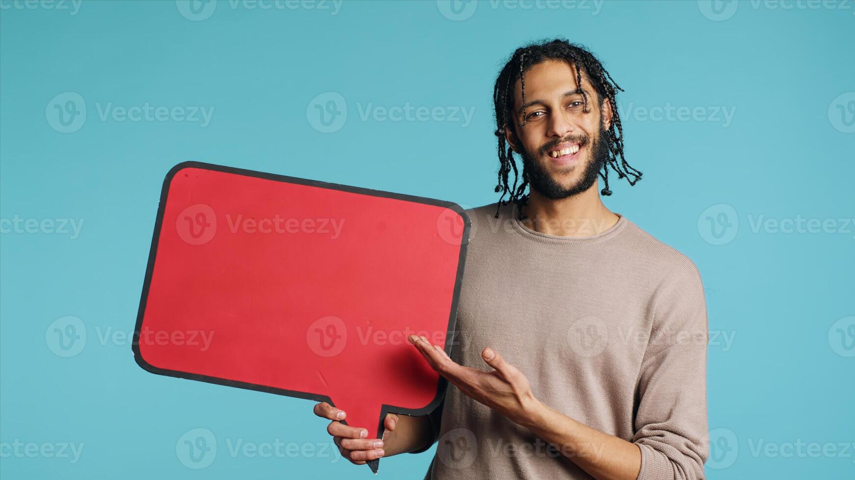 Happy man holding red speech bubble sign of empty space for text. Cheerful Middle Eastern person presenting thought bubble cardboard, isolated over blue studio background photo