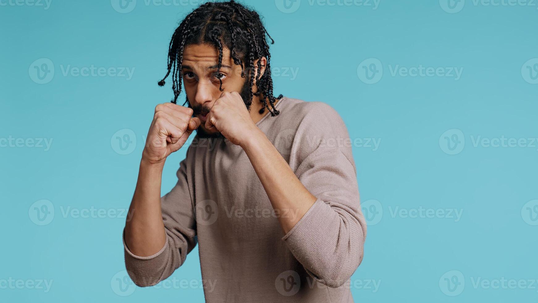Middle Eastern man pretending to fight, throwing punches, holding defensive guard. BIPOC martial artist person mock boxing, holding combat stance, exercising in blue studio background, camera B photo