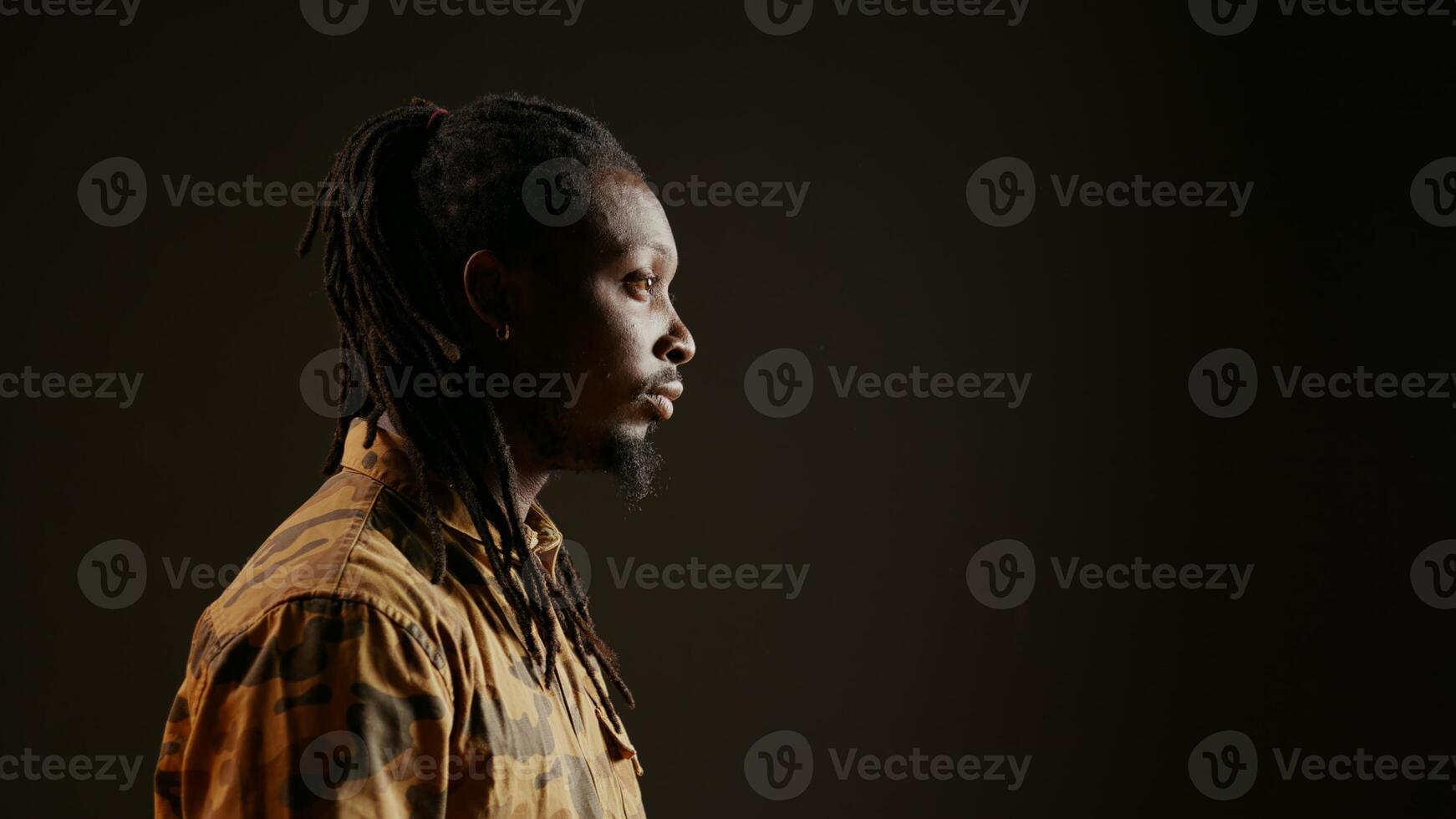 Smiling person posing in studio with black background, feeling confident wearing dreads and camo clothing. Cool guy with trendy hair acting stylish on camera, attractive man. Handheld shot. photo