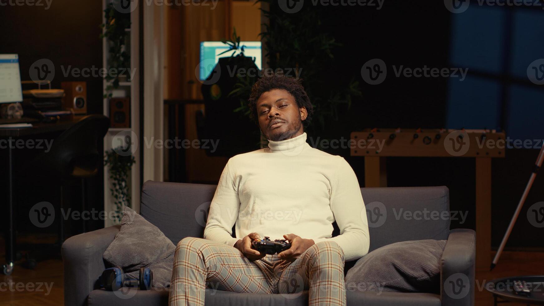 African american man in living room playing video games on gaming console from couch during leisure time. Gamer battling enemies in online multiplayer shooter from cozy apartment photo