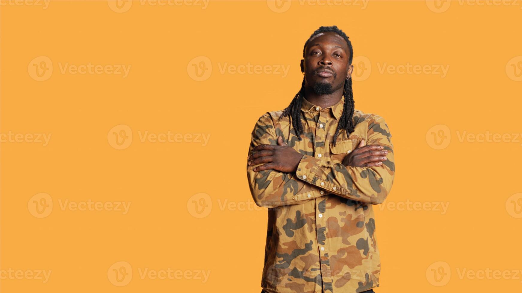 Portrait of guy with dreads smiling in studio set, wearing trendy camo clothes and feeling joyful on camera. African american adult with cool hair posing over orange background, natural style. photo