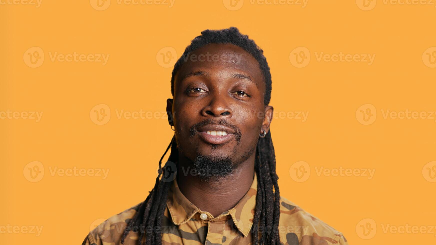 Confident person smiling in front of camera and posing, feeling trendy with hair braids and camo clothes in studio. Young adult being relaxed, cool trendy style over black backdrop. Handheld shot. photo