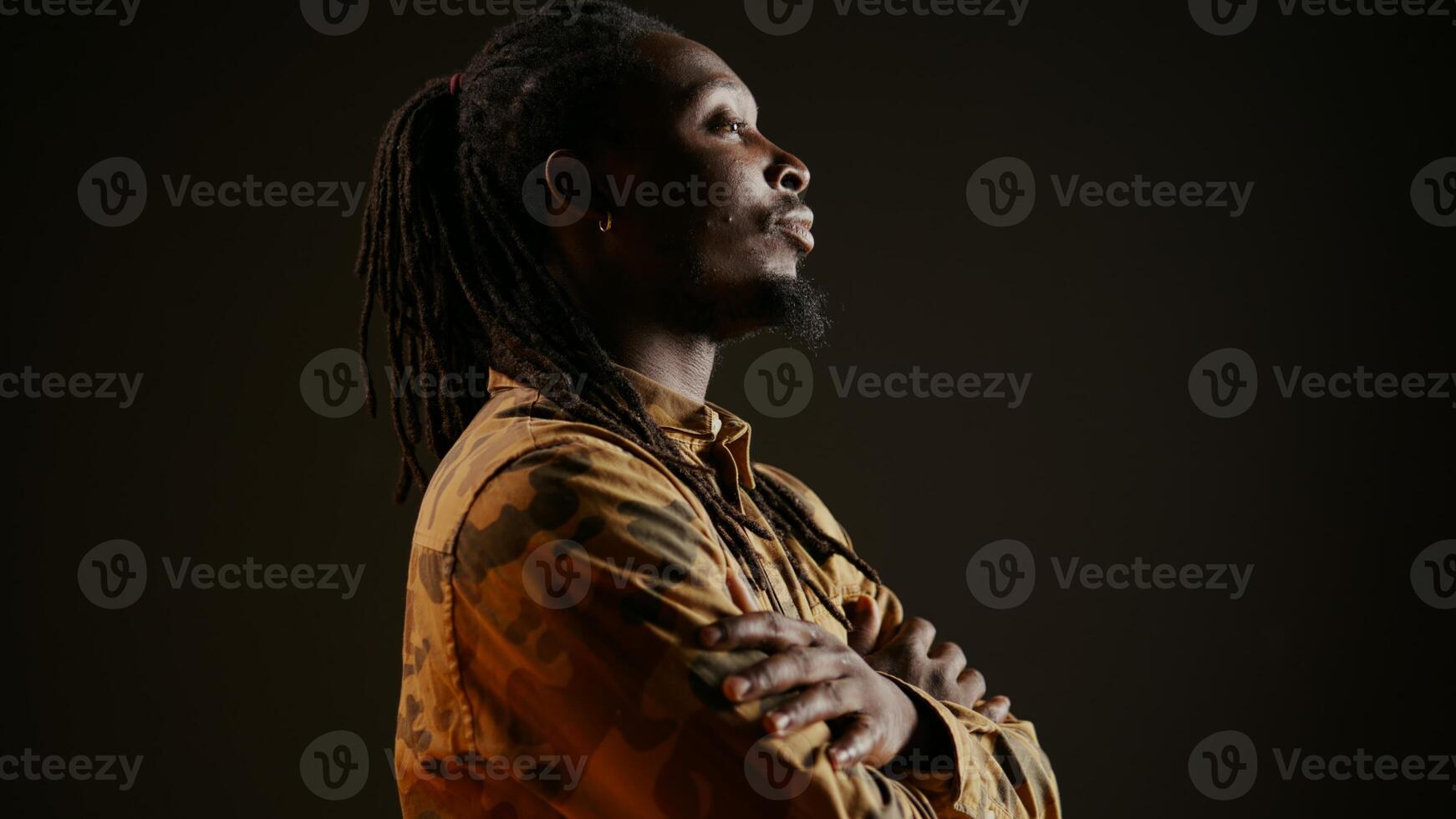 Trendy cool guy smiling in front of studio camera and posing, feeling confident with hair dreads and camo clothing items. Person being cheerful, modern fashion style over backdrop. Handheld shot. photo