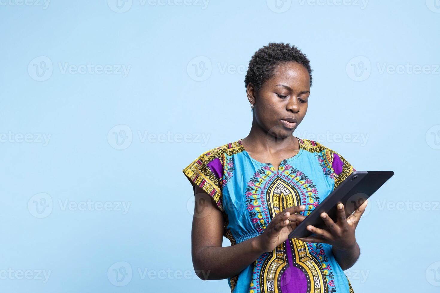 African american modern girl feels satisfied using a tablet over blue background, browsing online websites on mobile gadget. Young adult holds electronic device to navigate the internet. photo