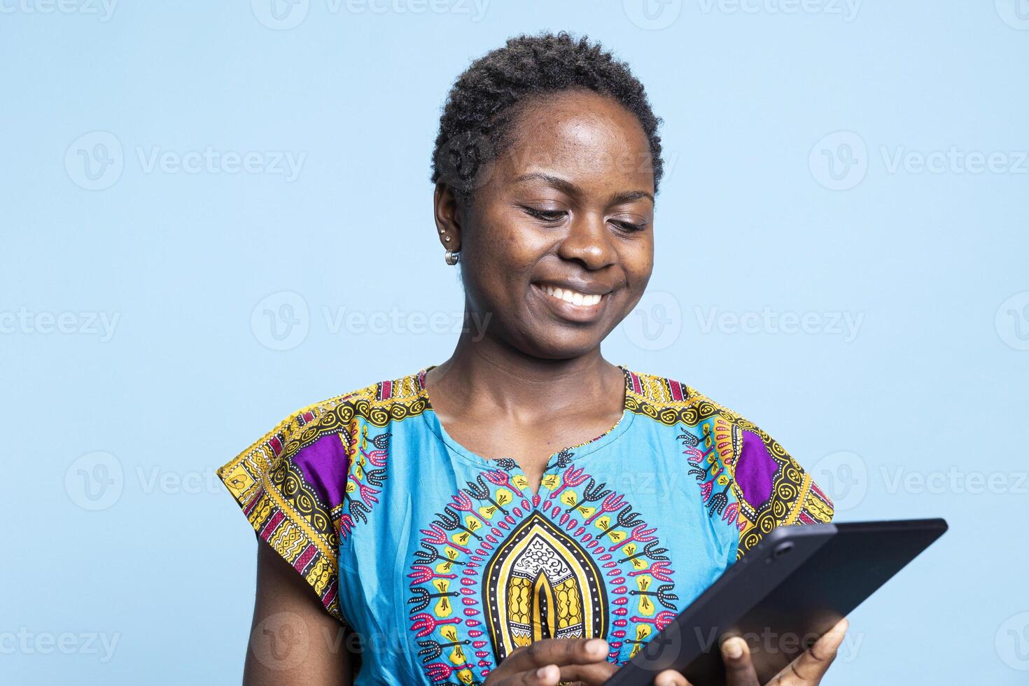 African american girl is pleased to use tablet against blue backdrop, access webpages on her mobile device. Using electronic gadget, young person searches the worldwide web in studio. photo