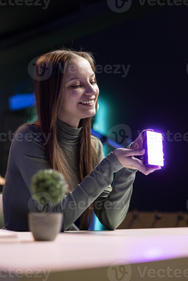 Happy young influencer in studio using camera to capture live broadcast, reviewing mini RGB lights. Joyful media star films electronics haul, presenting wireless LED lighting device photo