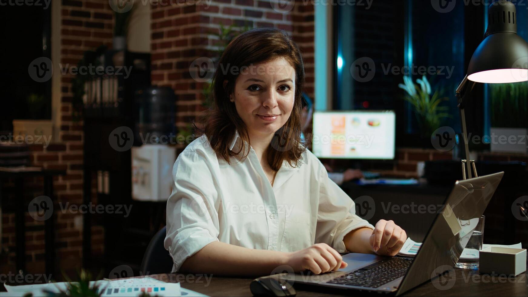 Portrait of smiling accountant researching key data for company project, doing tasks in office overnight. Happy employee sitting at desk in front of laptop, looking over accounting figures, camera A photo