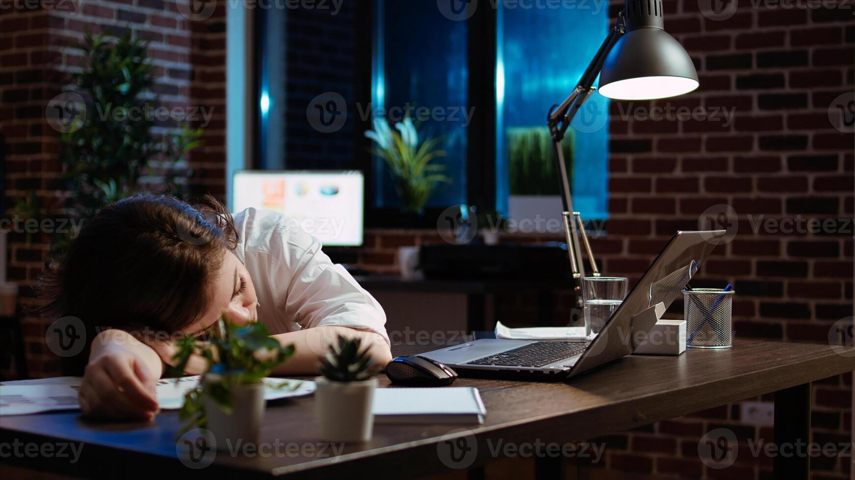 Sleepy employee asleep on computer desk late at night in brick wall office. Panning shot of tired businesswoman sleeping with head on table while working overnight on laptop, camera A photo