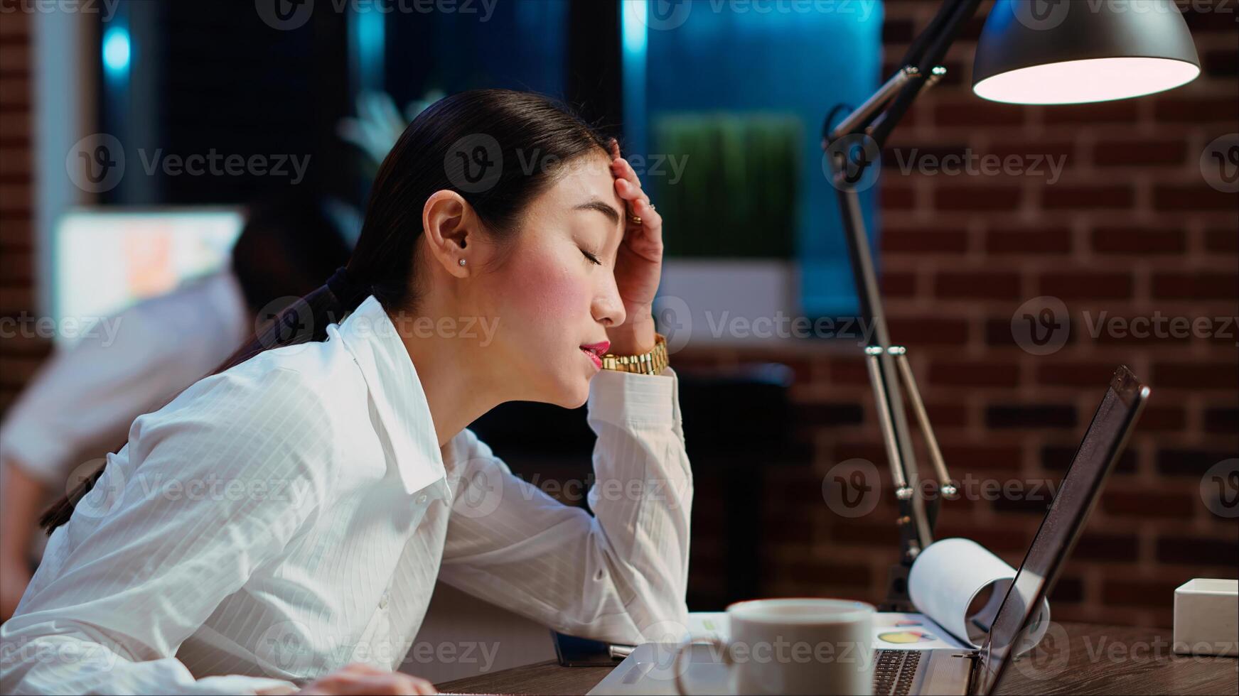 Bookkeeper feeling dread while having to do complicated tax reports in office at night. Worker squinting eyes in exasperation, feeling overworked while using laptop to do project research, camera B photo