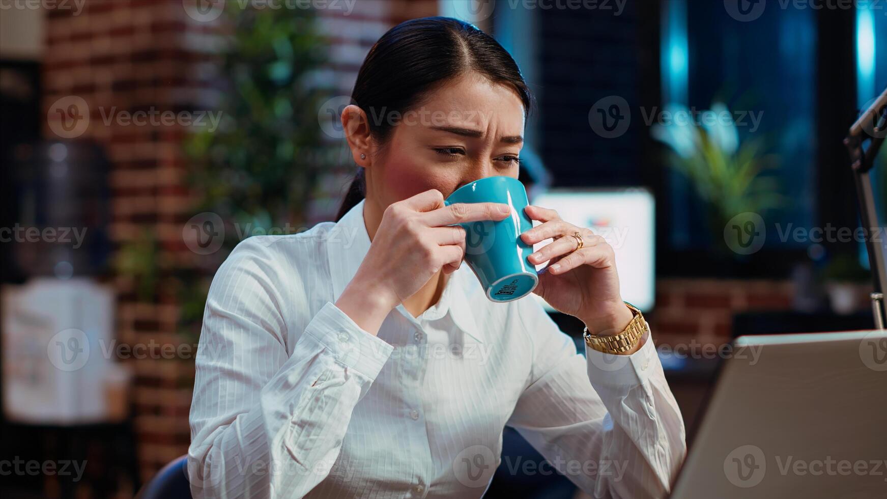 Employee tired while writing emails at computer desk, trying to freshen up by drinking coffee. Exhausted worker working overnight, typing on laptop, trying to get energy from caffeine, camera B photo