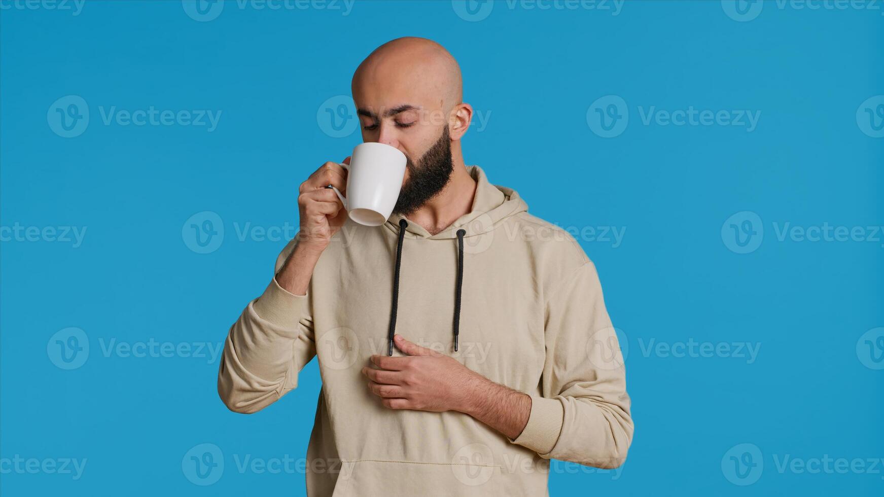 Muslim person enjoying fresh caffeine aroma in studio, drinking cup of coffee and standing over blue background. Middle eastern adult smelling beverage and taking a sip. Camera 1. photo