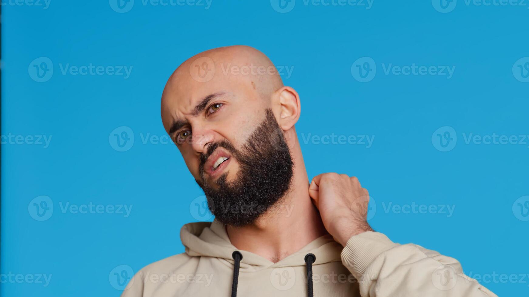 Sick middle eastern guy suffers from neck pain on camera, dealing with sore throat and muscle pressure over blue background. Adult being unwell with spine discomfort in studio. Camera 2. Handheld shot. photo