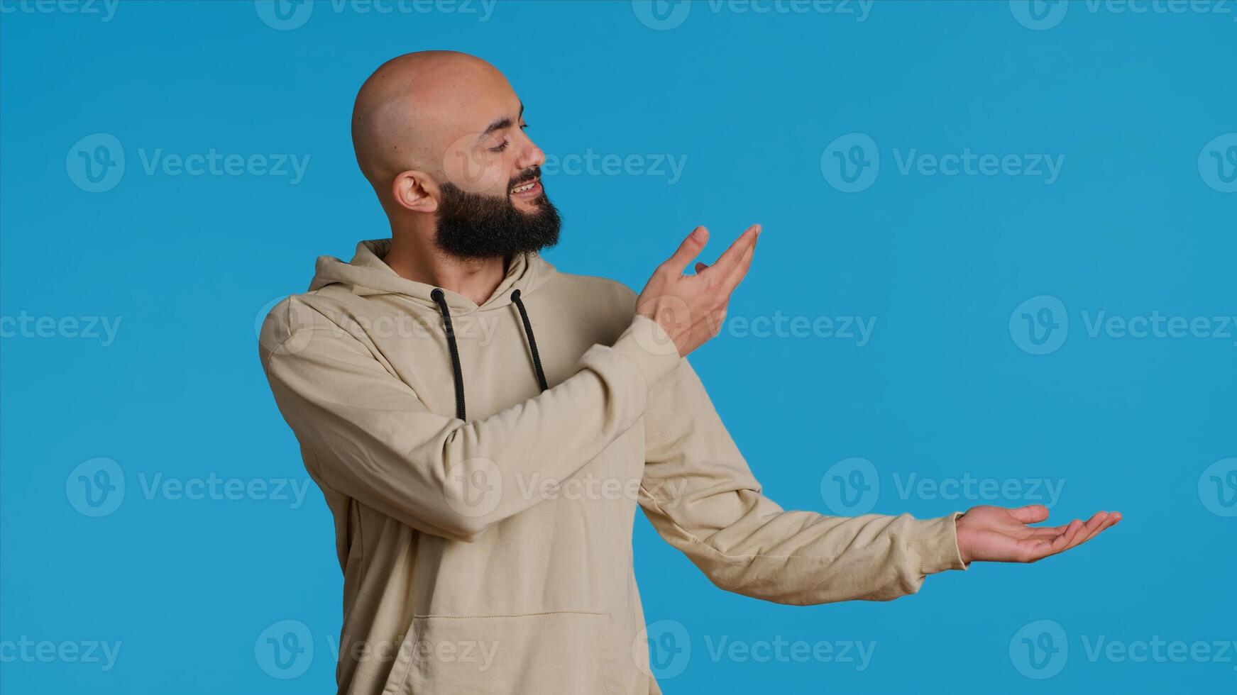 Cheerful man creating ad in front of studio camera, pointing to left or right sides over blue background. Middle eastern person showing commercial or advertisement for marketing campaign. Camera 1. photo