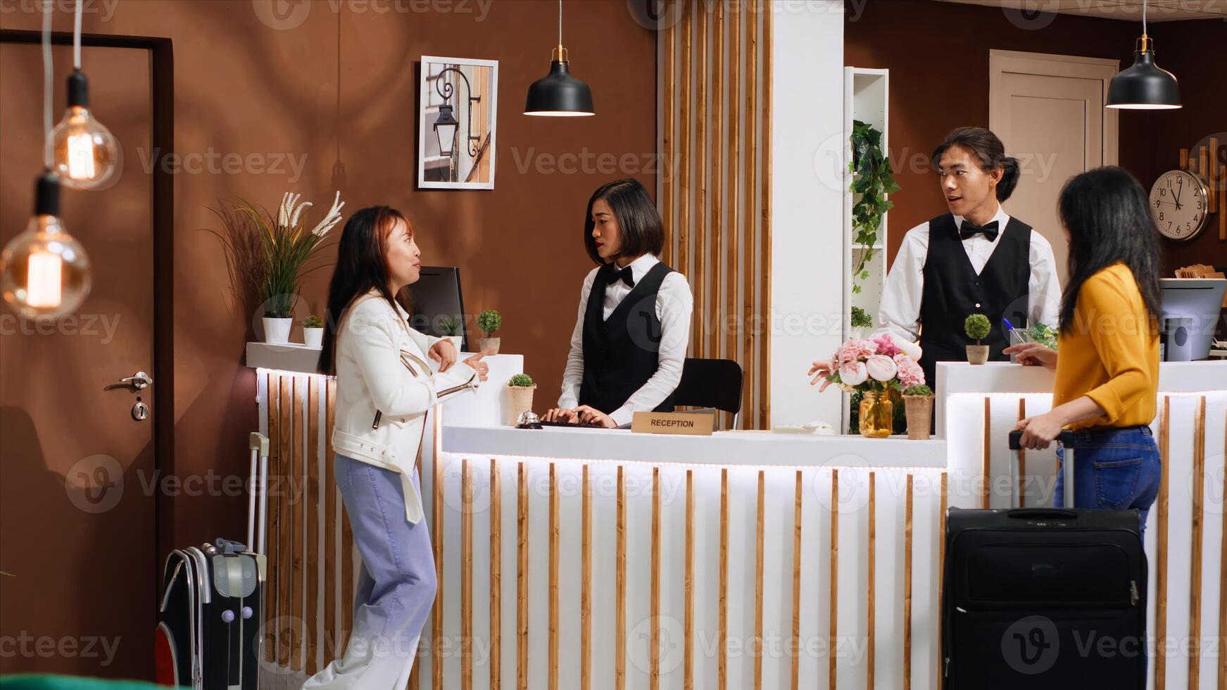 Asian friends arriving at hotel reception after travelling, waiting to check in and start city break vacation together. Two women guests receiving excellent concierge services from front desk staff. photo