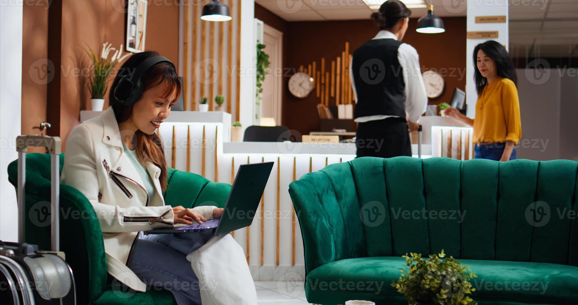 Asian woman enjoying movie on laptop in hotel lounge area, passing time waiting to be called at front desk. Young traveler using free wifi on computer, relaxing while she watches film. photo