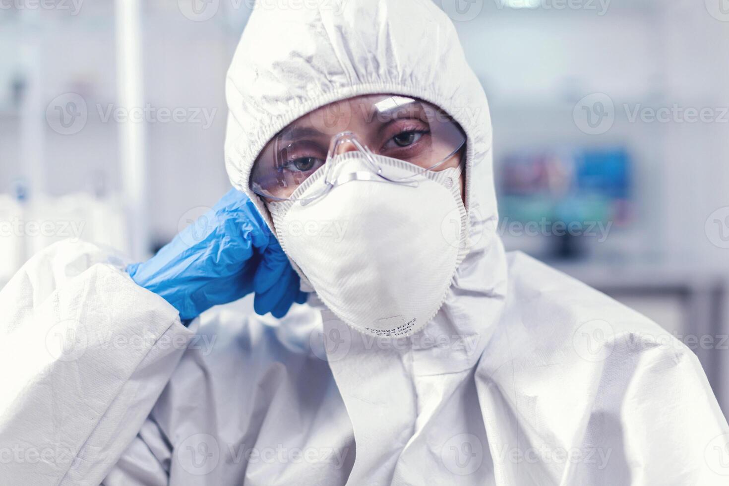 Portrait of overworked biolog looking at camera in modern equipped lab. Tired woman scientist in biotechnology laboratory wearing protective suit during global epidemic. photo