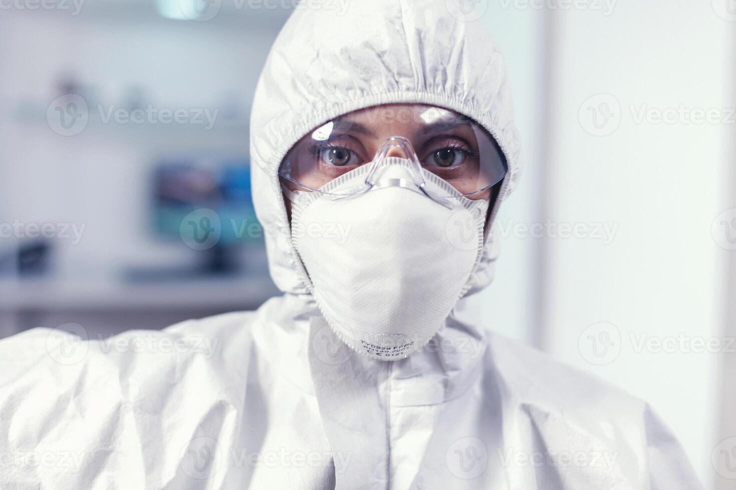 Exhausted microbiologist in coverall sitting in laboratory looking at camera. Tired woman scientist in biotechnology laboratory wearing protective suit during global epidemic. photo