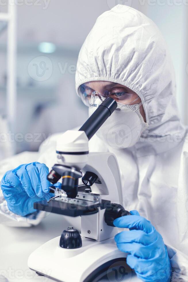 Woman scientis in coverall doing coronavirus investigation looking through microscope. Chemist researcher during global pandemic with covid-19 checking sample in biochemistry lab photo