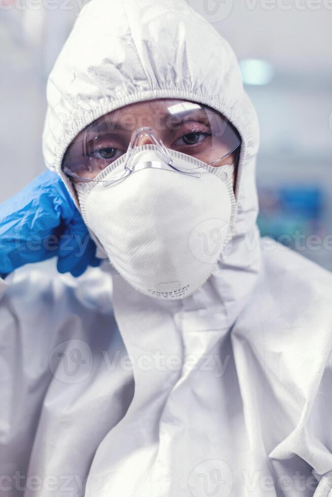 Tired laboratory doctor with overall suit looking at camera in equipped lab. Overworked woman scientist in biotechnology laboratory wearing protective suit during global epidemic. photo