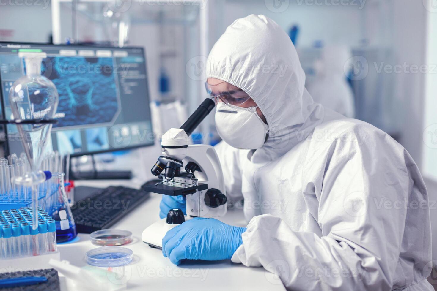 Biochemist wearing protective suit working in lab with modern microscope. Virolog in coverall during coronavirus outbreak conducting healthcare scientific analysis. photo