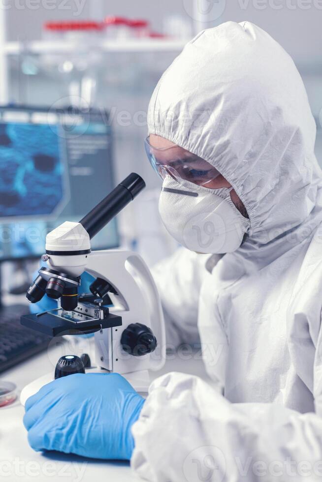 Close up of chief researcher scientist adjusts microscope in time of coronavirus experiment. Virolog in coverall during coronavirus outbreak conducting healthcare scientific analysis. photo