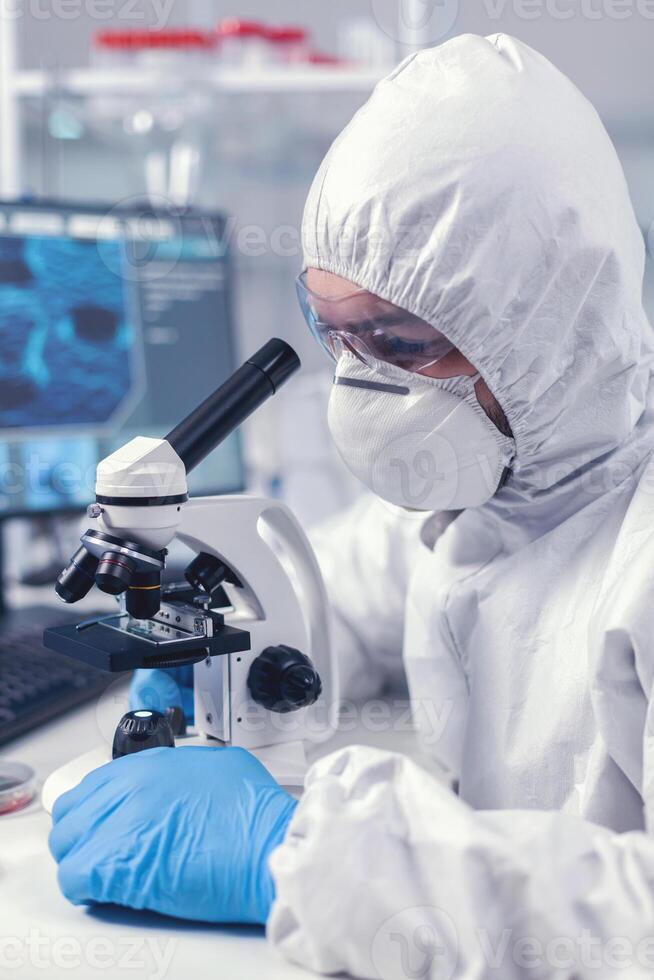 joven científico Mira mediante microscopio mientras haciendo investigación en científico laboratorio vestido en ppe traje. virologo en bata durante coronavirus brote conductible cuidado de la salud científico análisis. foto