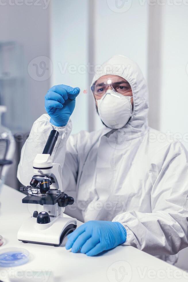 Microbiologist dressed in ppe suit analyzing sample on microscope glass slide Examining vaccine evolution using high tech for researching treatment against covid19 virus. photo