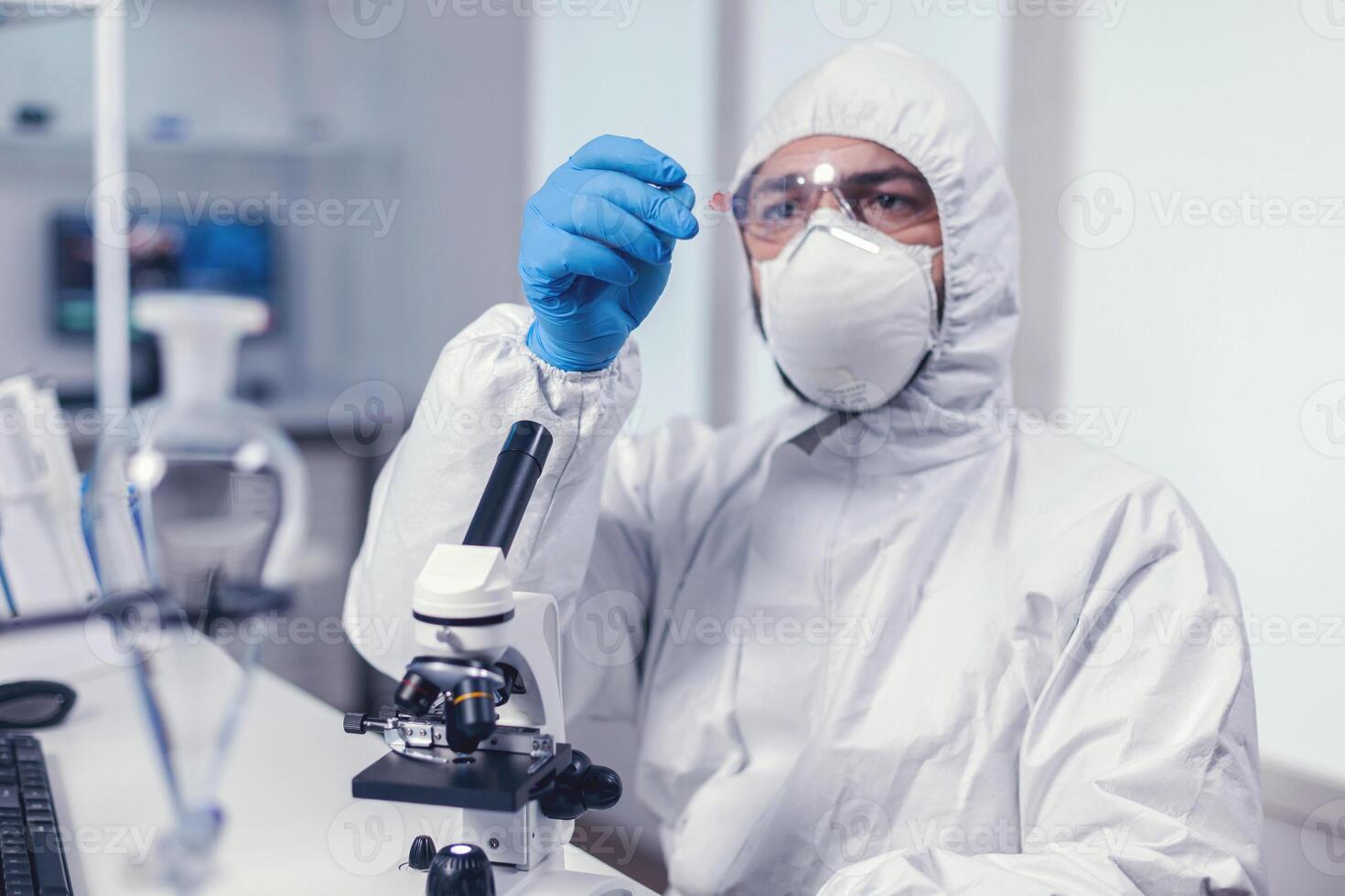 Scientist looking at a microscope slide dressed in coverall during coronavirus. Examining vaccine evolution using high tech for researching treatment against covid19 virus. photo