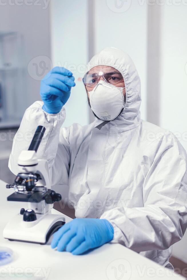 Medical engineer looking attentive at sample on glass slide sitting at his workplace dressed in ppe suit. Examining vaccine evolution using high tech for researching treatment against covid19 virus. photo