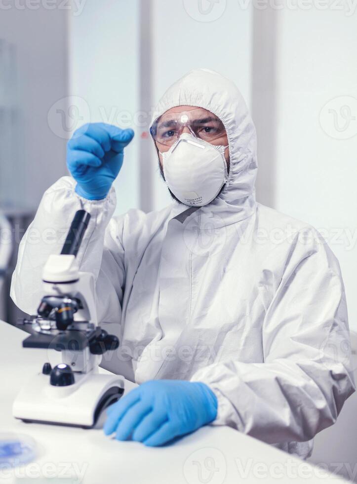 Medical chemist holding microscop glass slide with blood sample dressed in ppe suit in modern lab.. Examining vaccine evolution using high tech for researching treatment against covid19 virus. photo