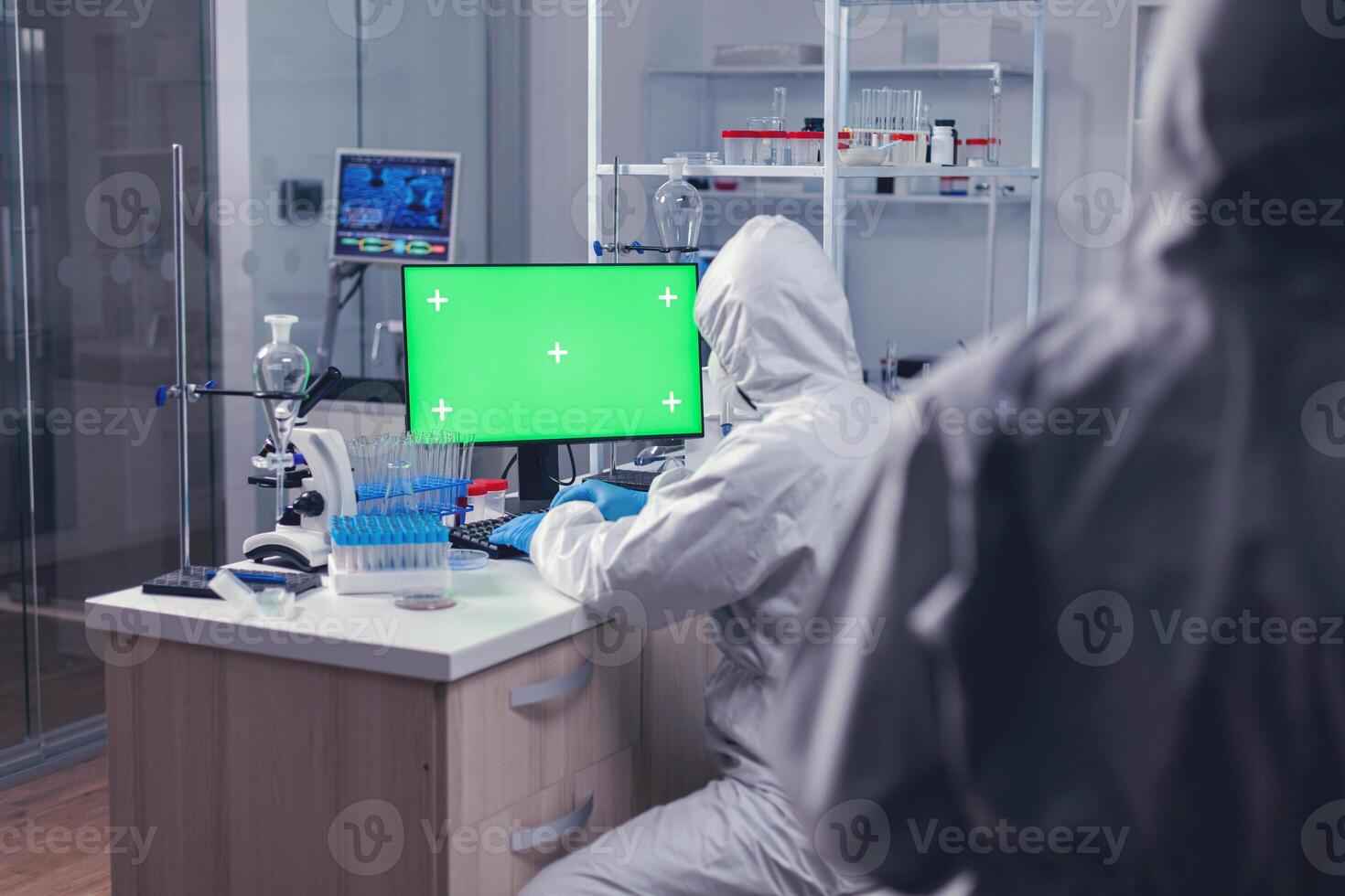 Scientist dressed in ppe suit working on computer with green screen during global pandemic. Team of microbiologists doing vaccine research writing on device with chroma key, isolated, mockup display. photo