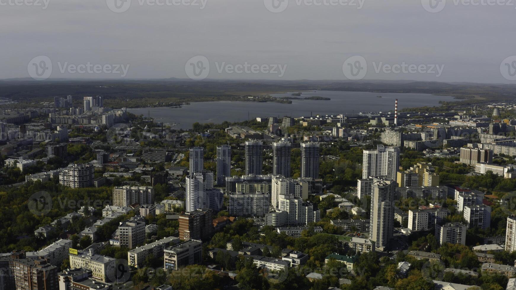 City with high-rise buildings on background of lake and green forest. Stock footage. Modern buildings in beautiful green city with lake and forest horizon. Top view of panorama of modern city in photo