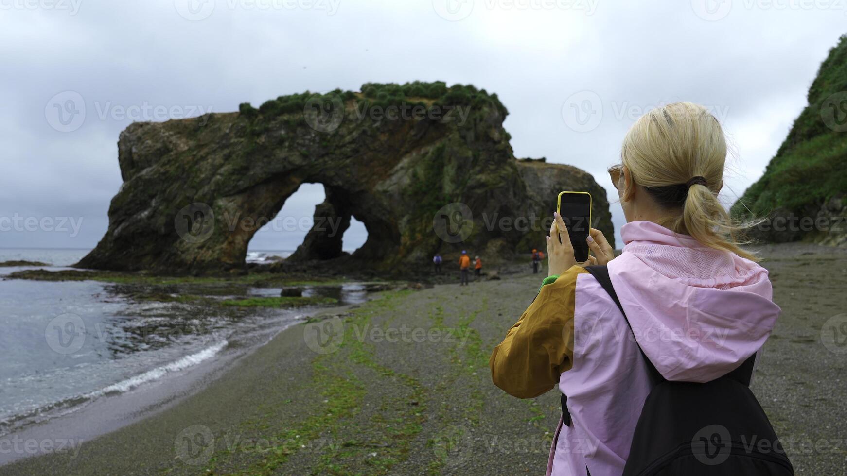 Woman on shore takes pictures of sea on phone. Clip. Woman photographs seashore with rocky arch. Group of tourists takes pictures of rocky arch on seashore photo