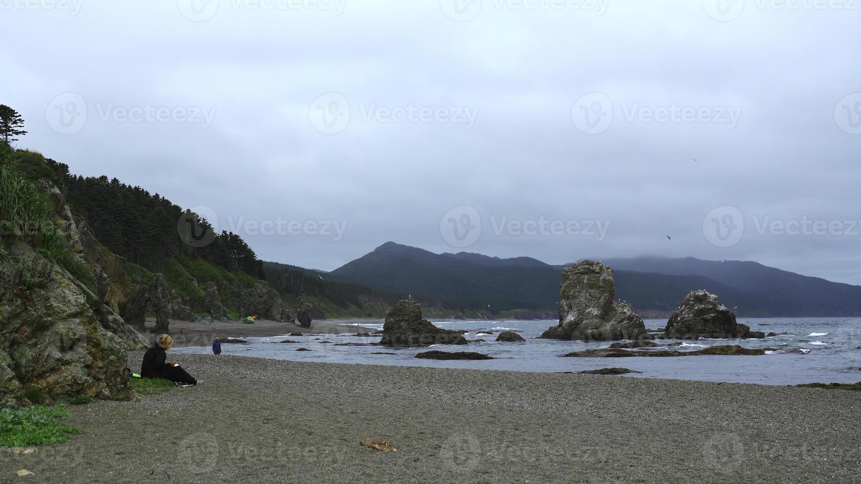 People relax on rocky coast with mountains on cloudy day. Clip. Beautiful coast with rocks and rocks sticking out on shore. People relax on beautiful coast with rocks and mountains photo